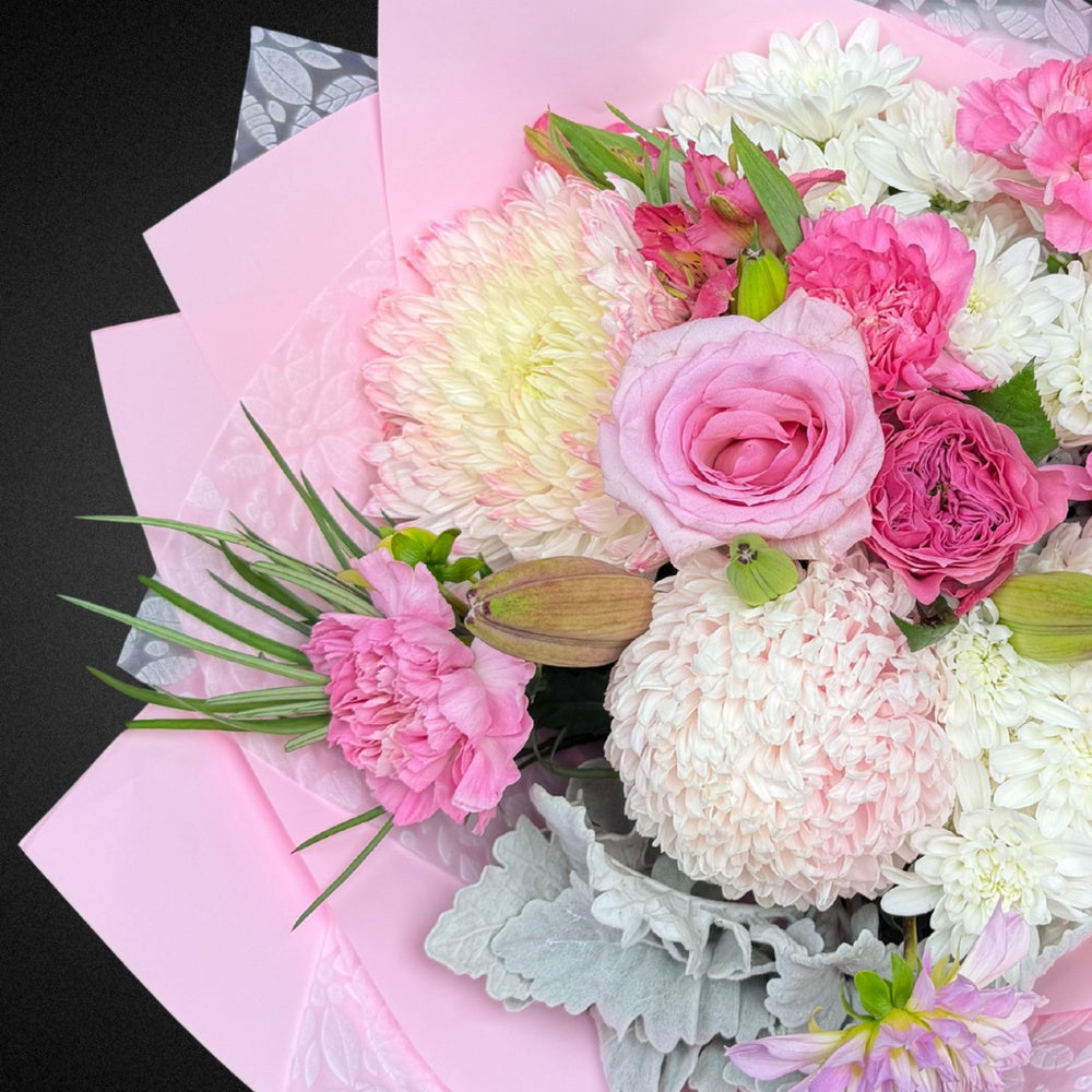Close-up of a premium Mother's Day bouquet showcasing delicate pastel pink roses, cheerful white chrysanthemums, soft peach carnations, and silver-grey Dusty Miller leaves, highlighting the mixed pastel palette and fresh blooms.