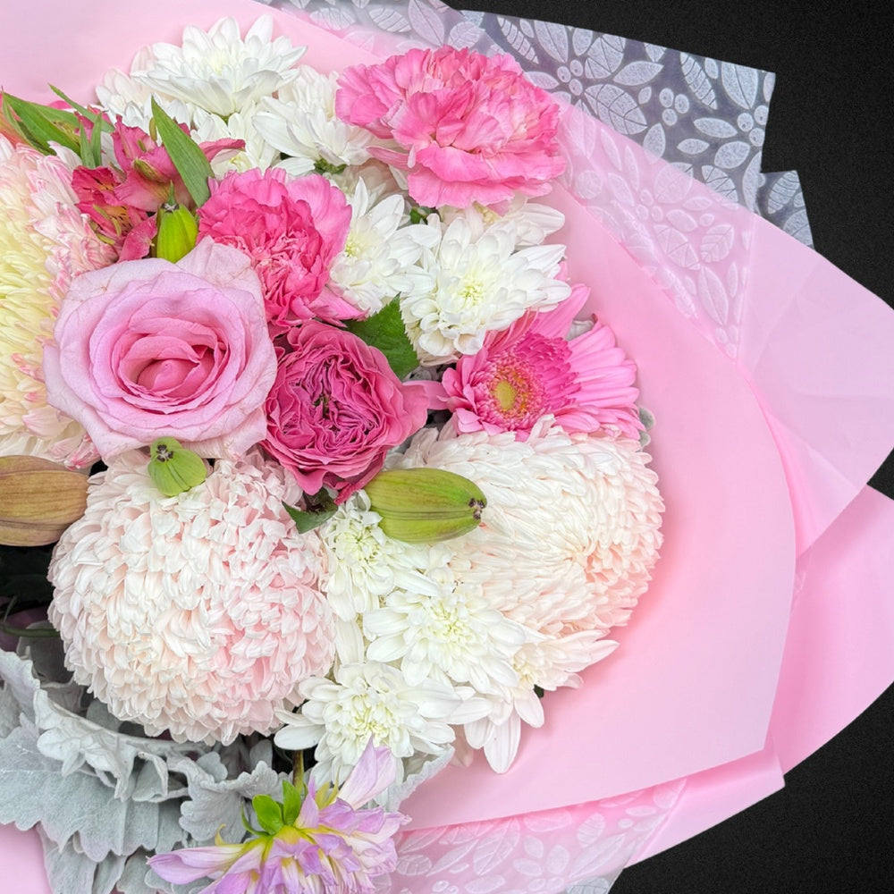 Close-up of a premium Mother's Day bouquet showcasing delicate pastel pink roses, cheerful white chrysanthemums, soft peach carnations, and silver-grey Dusty Miller leaves, highlighting the mixed pastel palette and fresh blooms.