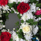 Close-up of a handcrafted wreath showcasing delicate pink roses, yellow chrysanthemums, and red carnations with lush green foliage.